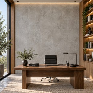 A modern home office setup featuring a large rectangular wooden desk as the central product, styled with a black office chair, a black desk lamp, a small vase with greenery, and books. The desk is set on a textured beige rug against a concrete-textured wall, with a large window on the left and built-in wooden shelves with plants on the right.